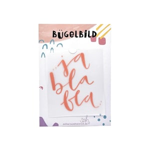 Puede incluir: Una lámina de vinilo de transferencia térmica con las palabras "ja bla bla" en escritura coral. Las palabras están sobre un fondo transparente, con la palabra "BÜGELBILD" en la parte superior. La lámina está sobre un fondo blanco con elementos decorativos.