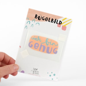 Op de afbeelding: Een witte kaart met een roze en geel ontwerp. De kaart zegt "Bügelbild" en heeft een sticker met de tekst "ich bin genug" in paarse en turquoise letters op een perzikkleurige achtergrond.