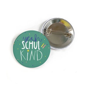 Op de afbeelding: Een groene knop met de tekst "yeah schul kind" in blauwe en gele letters. De knop heeft een witte achtergrond met een rasterpatroon en kleurrijk confetti. De achterkant van de knop is zilver met een pin.