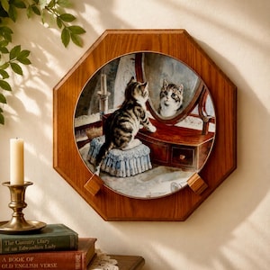 May include: A decorative wall plate featuring a circular illustration of a tabby kitten sitting on a blue tufted stool, looking into a wooden vanity mirror. The plate is mounted within an octagonal wooden frame with a warm brown finish.