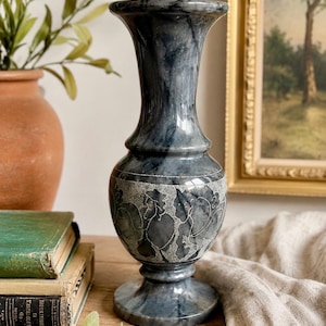 May include: A tall, polished dark gray marble vase featuring a smooth, flared rim and a rounded body. The central section of the vase is decorated with a wide, textured band displaying etched floral patterns in a lighter gray stone finish.
