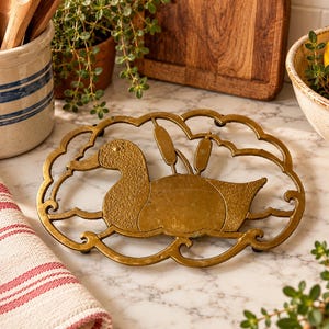 May include: An oval-shaped brass trivet featuring a textured duck silhouette swimming among stylized cattails. The metal piece has an openwork border with swirling wave-like patterns and a warm, metallic gold finish with visible patina.