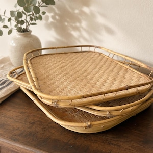 May include: A set of three rectangular woven rattan trays with rounded corners and raised edges. The trays feature a light tan, natural bamboo-like finish with a tight, intricate basket-weave pattern on the base and a sturdy frame structure.