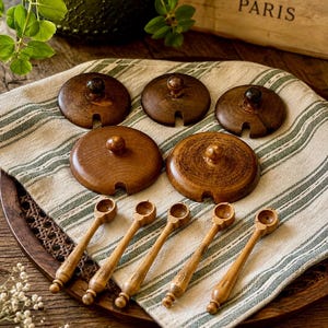 May include: A set of five circular dark brown wooden lids with central knobs and small notches, accompanied by five matching slender wooden spoons with rounded bowls, arranged on a beige and green striped fabric cloth.