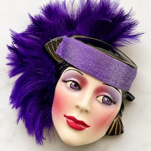 May include: A ceramic face mask with a purple feather headdress and a matching headband. The face has red lips, rosy cheeks, and purple eye makeup. The mask has a vintage, theatrical aesthetic.