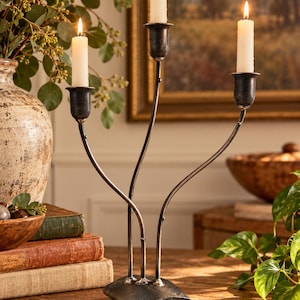 May include: A dark gray metal candelabra with three curved arms, each holding a white taper candle. The base is a solid, slightly arched metal piece. The structure features thin, branch-like stems that rise from the center to support the candle cups.