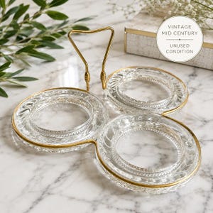 May include: A vintage mid-century three-part serving dish. The clear glass dishes have a gold rim and are connected by a gold handle. The text "VINTAGE MID CENTURY UNUSED CONDITION" is in a white circle.