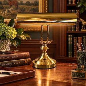May include: A polished brass banker lamp featuring a horizontal cylindrical shade and an adjustable arm, mounted on a circular base. The lamp emits a warm light, illuminating the surrounding desk area with a reflective metallic finish.