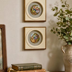 May include: Two square wall frames with cream-colored borders and circular centers, each containing a small, colorful circular plaque depicting a scene with figures in a landscape. The frames are mounted vertically one above the other on a wall.