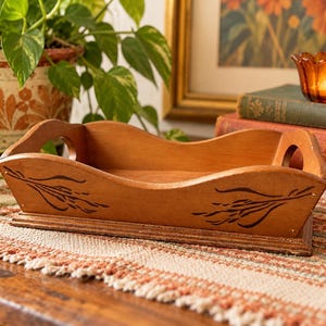 May include: A rectangular wooden tray with a light brown finish, featuring curved side handles and dark brown painted wheat-like botanical motifs on the front panel. The tray has a raised base and a smooth, open-top design.
