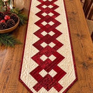 May include: A long, rectangular quilted table runner featuring a central geometric pattern of interlocking deep red and patterned burgundy fabric strips set against a cream-colored background with a diamond-stitched texture and red border.