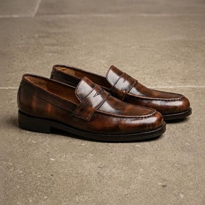 Dark brown leather penny loafers with glossy finish, classic strap detail, moccasin stitching, low stacked heels, and tan leather lining.