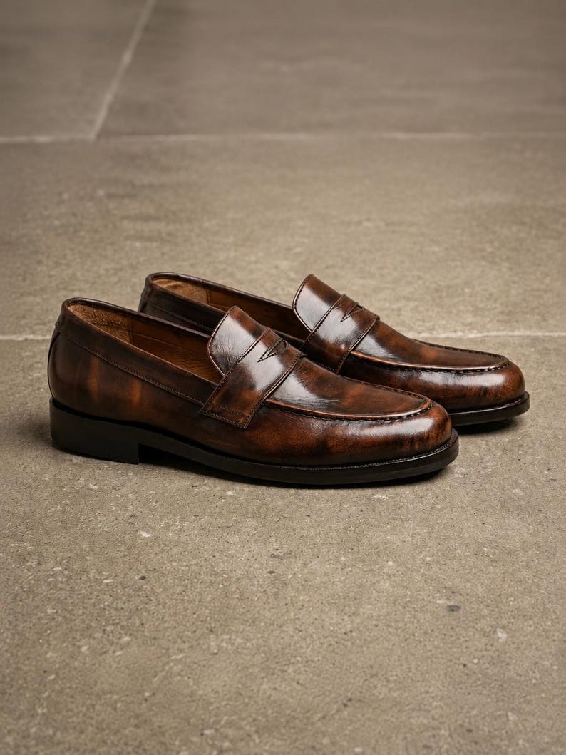 Dark brown leather penny loafers with glossy finish, classic strap detail, moccasin stitching, low stacked heels, and tan leather lining.