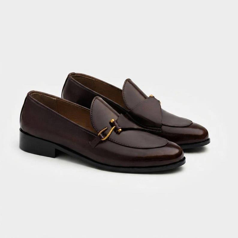 May include: A pair of dark brown leather loafers featuring a smooth, polished finish, a rounded toe, and a gold-toned metal horsebit detail across the vamp. The shoes have a low, stacked black heel and a structured silhouette with visible stitching.