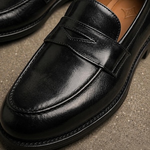Glossy black leather penny loafer close-up featuring a classic strap, moccasin stitching, polished finish, and tan leather lining.