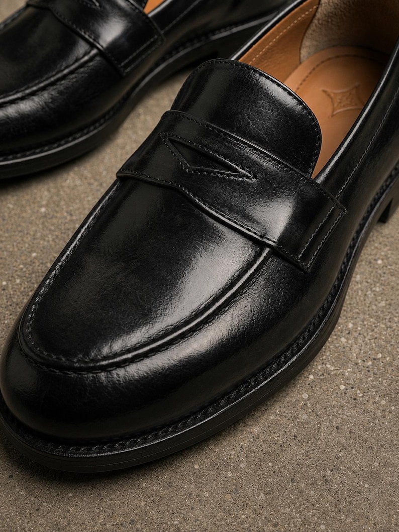 Glossy black leather penny loafer close-up featuring a classic strap, moccasin stitching, polished finish, and tan leather lining.