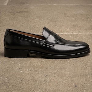 Black leather penny loafer shown from the side, featuring a glossy finish, classic strap detail, moccasin stitching, and a low stacked heel.