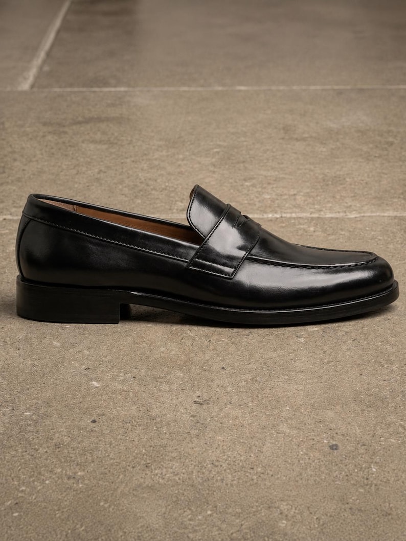 Black leather penny loafer shown from the side, featuring a glossy finish, classic strap detail, moccasin stitching, and a low stacked heel.