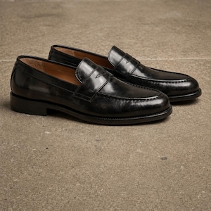 Black leather penny loafers with glossy finish, moccasin toe stitching, slip-on design, and classic leather soles on a textured surface.