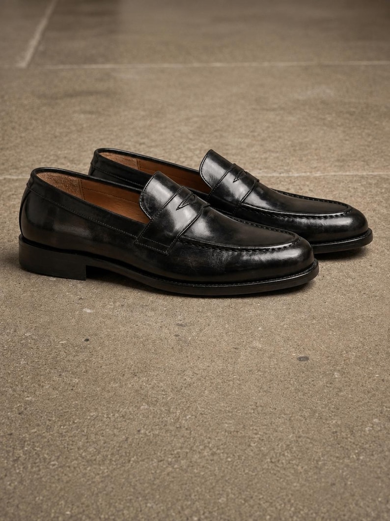 Black leather penny loafers with glossy finish, moccasin toe stitching, slip-on design, and classic leather soles on a textured surface.