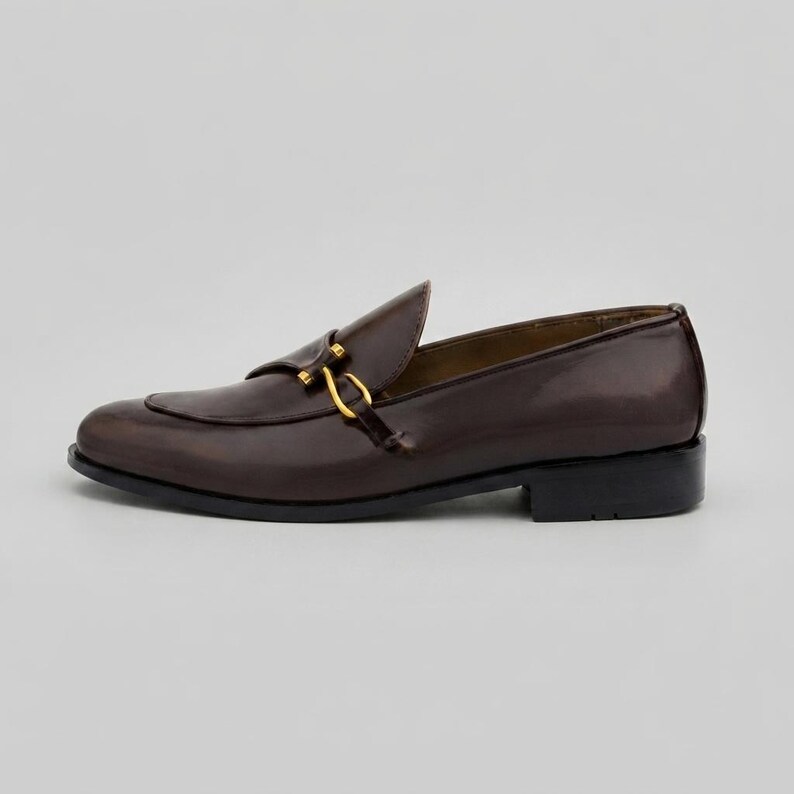 May include: A dark brown leather loafer featuring a polished finish and a gold-toned metal horsebit detail across the vamp. The shoe has a low block heel, a rounded toe, and a smooth, structured silhouette with visible stitching along the apron.