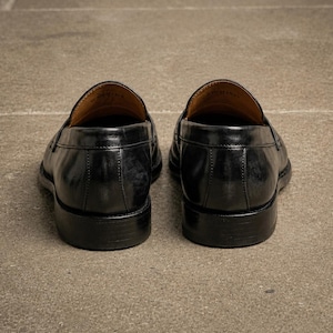 Black leather penny loafers viewed from the back, showing stitched heel panels, polished finish, tan leather lining, and stacked black heels.