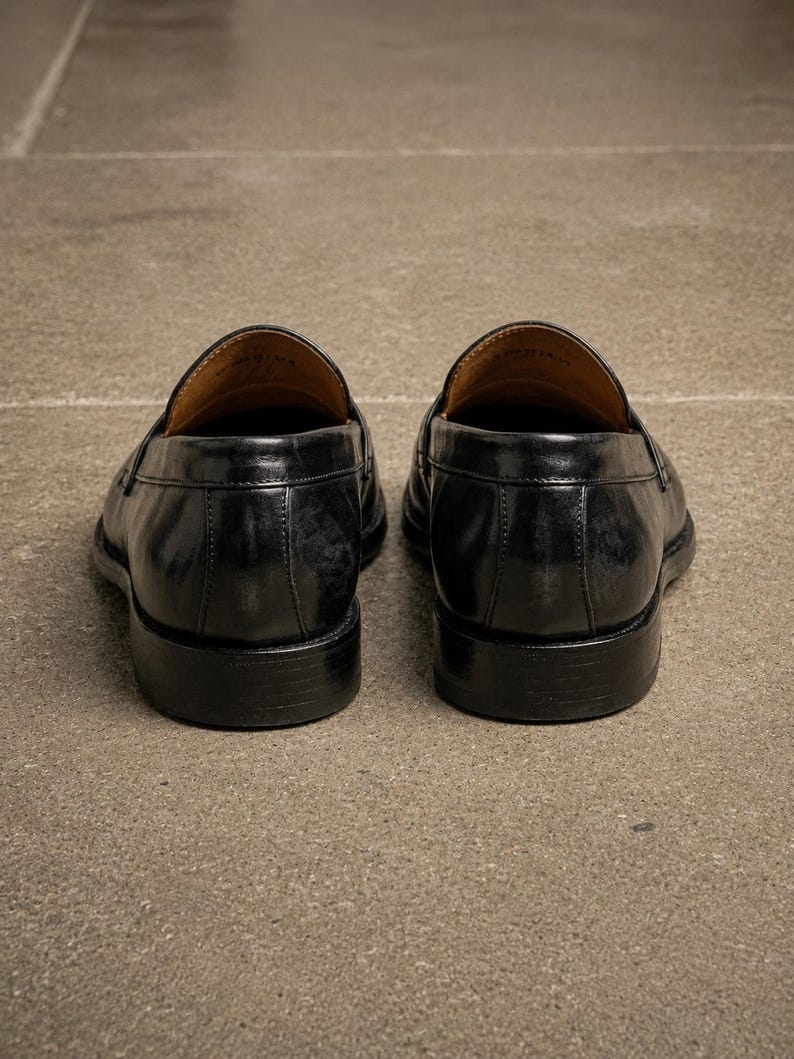 Black leather penny loafers viewed from the back, showing stitched heel panels, polished finish, tan leather lining, and stacked black heels.
