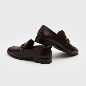 May include: A pair of dark brown leather loafers featuring a gold-toned metal horsebit detail across the vamp. The shoes have a smooth finish, a rounded toe, and a low black stacked heel, presented against a plain white background.