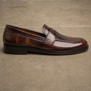 Dark brown leather penny loafer in side profile, featuring a glossy finish, classic strap detail, moccasin stitching, and a low stacked heel.