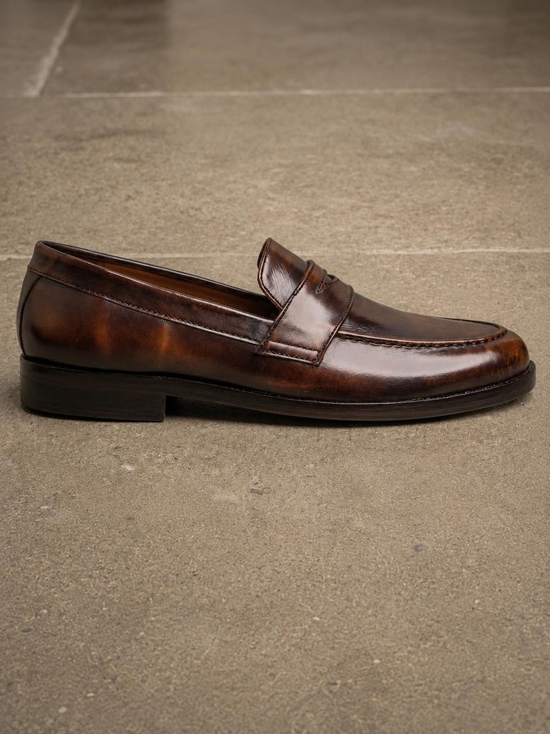 Dark brown leather penny loafer in side profile, featuring a glossy finish, classic strap detail, moccasin stitching, and a low stacked heel.