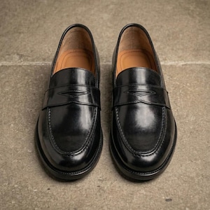 Black leather penny loafers with a glossy finish, tan leather interiors, and classic stitched detailing, photographed from above on a textured gray concrete surface.