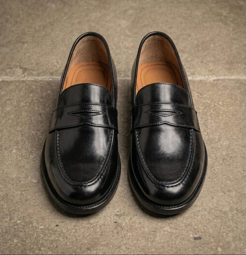 Black leather penny loafers with a glossy finish, tan leather interiors, and classic stitched detailing, photographed from above on a textured gray concrete surface.
