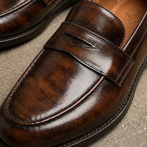 Close-up of a dark brown leather penny loafer showing polished finish, classic strap detail, moccasin toe stitching, and tan leather lining.