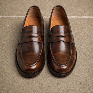 Polished dark brown leather penny loafers with classic strap detail, moccasin toe stitching, glossy finish, and tan leather lining.