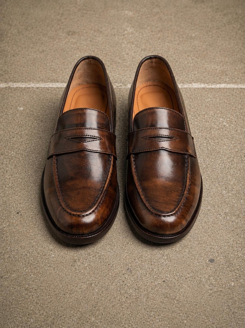 Polished dark brown leather penny loafers with classic strap detail, moccasin toe stitching, glossy finish, and tan leather lining.