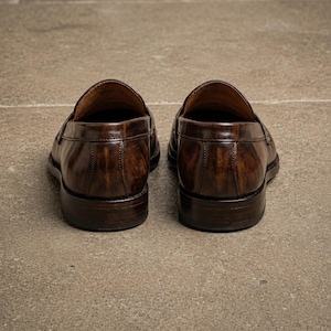 Dark brown leather penny loafers viewed from the back, showing polished heel panels, vertical stitching, low stacked heels, and leather lining.