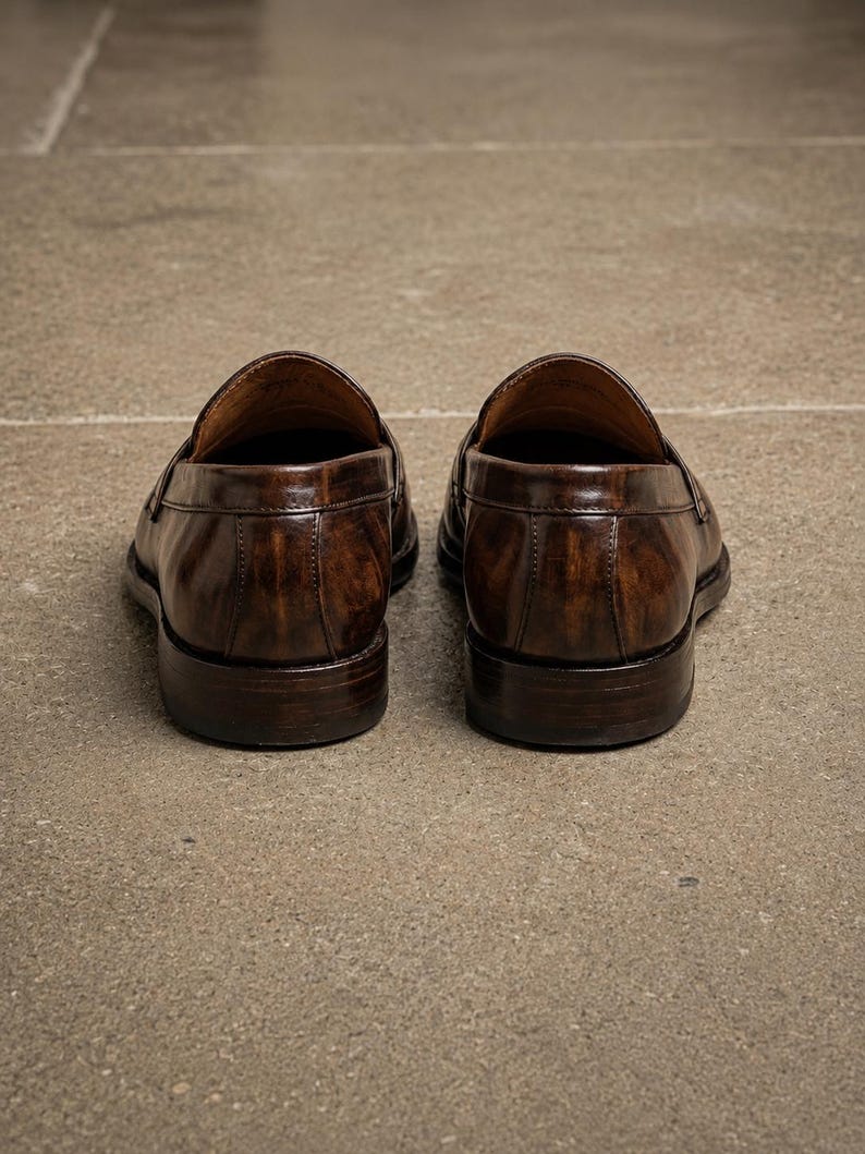 Dark brown leather penny loafers viewed from the back, showing polished heel panels, vertical stitching, low stacked heels, and leather lining.