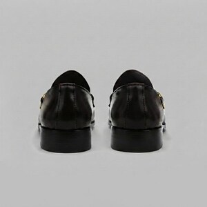 May include: A pair of black leather loafers shown from the back view. The shoes feature a smooth, polished finish, a low stacked heel, and a gold-toned metal horsebit detail visible on the side of each shoe. The design is simple and structured.