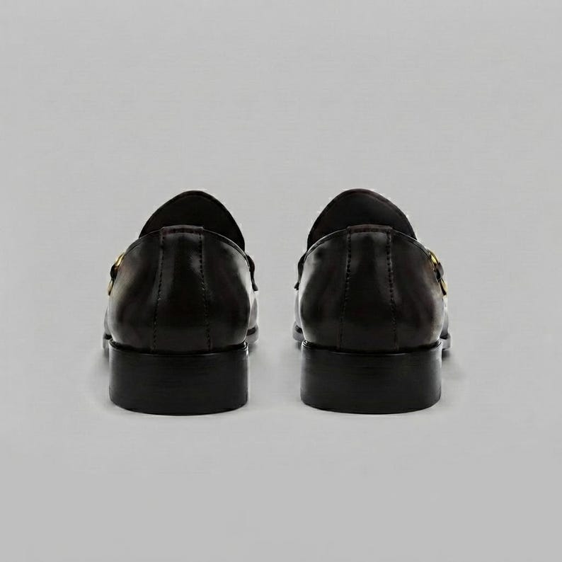 May include: A pair of black leather loafers shown from the back view. The shoes feature a smooth, polished finish, a low stacked heel, and a gold-toned metal horsebit detail visible on the side of each shoe. The design is simple and structured.