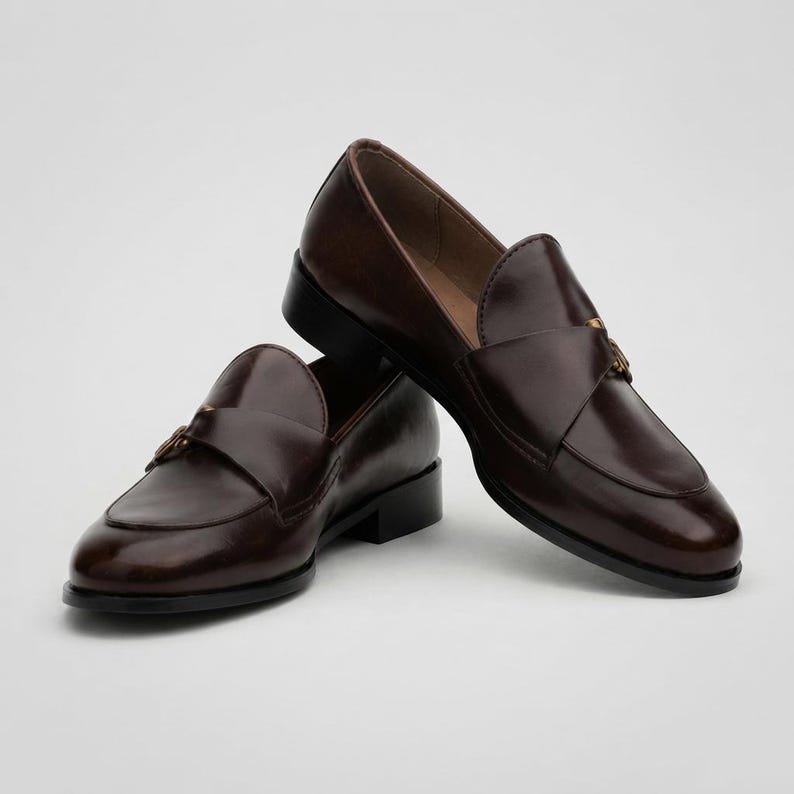 May include: A pair of dark brown leather loafers featuring a smooth finish, a rounded toe, and a decorative strap across the vamp with a small metallic buckle detail. The shoes have a low block heel and a structured silhouette with visible stitching.