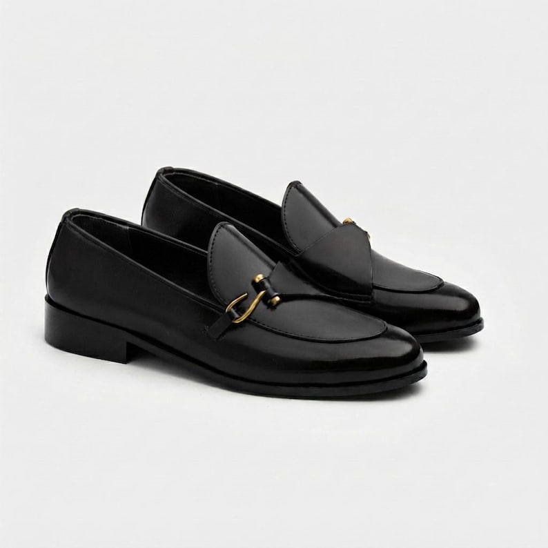 May include: A pair of black loafers with a polished finish, featuring a rounded toe and a gold-toned metal horsebit detail across the vamp. These slip-on shoes have a low block heel and a structured silhouette with visible stitching along the edge.