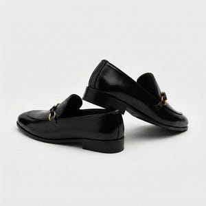 May include: A pair of black leather loafers featuring a polished finish and a gold-toned metal horsebit detail across the vamp. The shoes have a low block heel, a rounded toe, and a structured silhouette, presented in a side-profile view.
