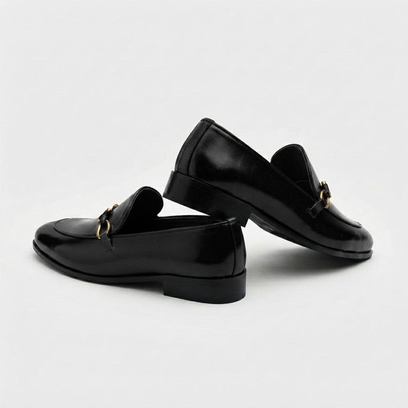 May include: A pair of black leather loafers featuring a polished finish and a gold-toned metal horsebit detail across the vamp. The shoes have a low block heel, a rounded toe, and a structured silhouette, presented in a side-profile view.