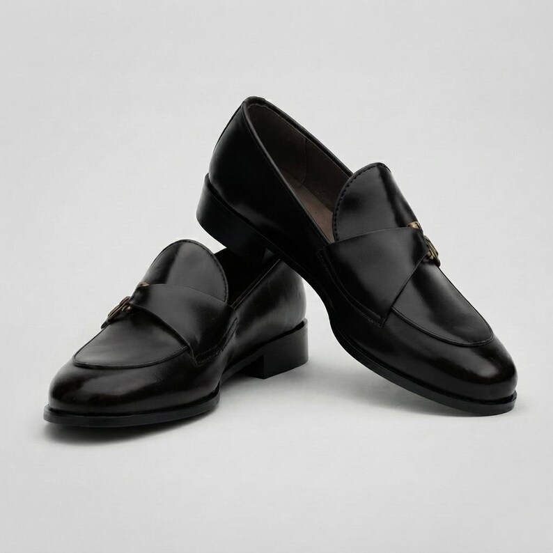 May include: A pair of black leather loafers featuring a smooth, polished finish, a rounded toe, and a decorative strap across the vamp secured with a small metallic buckle detail on the side. The shoes have a low block heel and a structured silhouette.