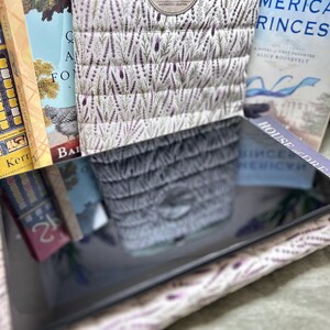 May include: A quilted fabric cover featuring a lavender and green botanical pattern with a circular Mallard Collective logo. The cover is positioned in front of several books, including titles like American Princess and House of Dreams, with a reflective surface.