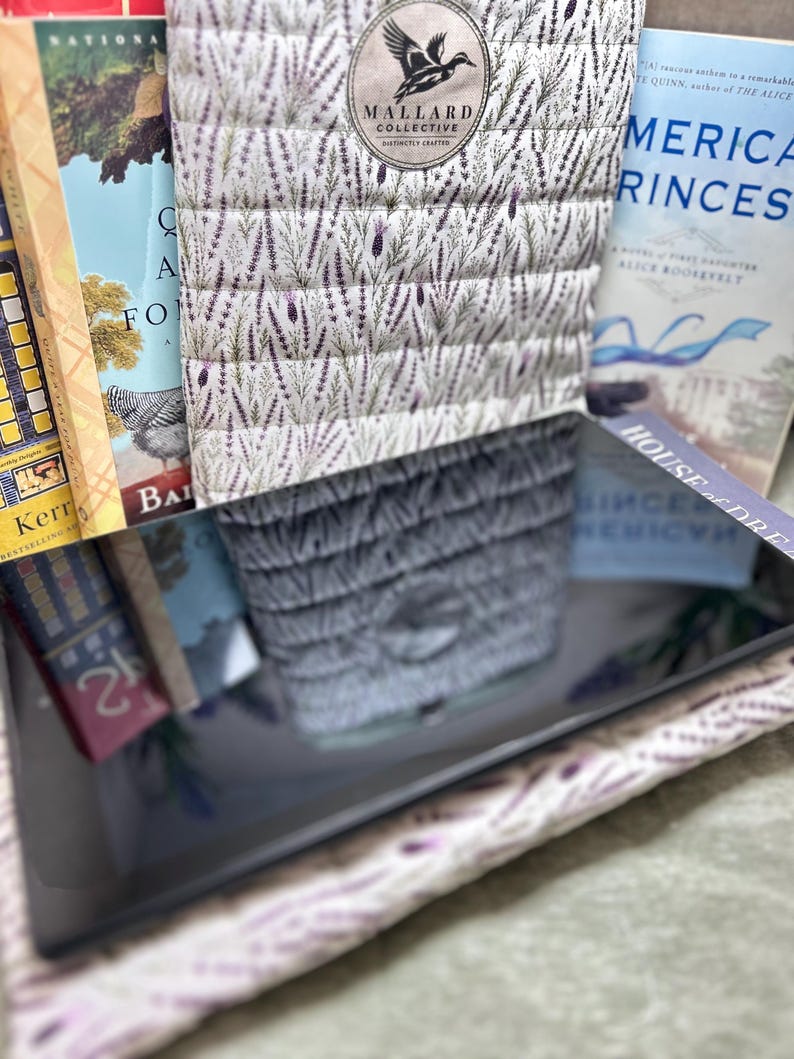 May include: A quilted fabric cover featuring a lavender and green botanical pattern with a circular Mallard Collective logo. The cover is positioned in front of several books, including titles like American Princess and House of Dreams, with a reflective surface.