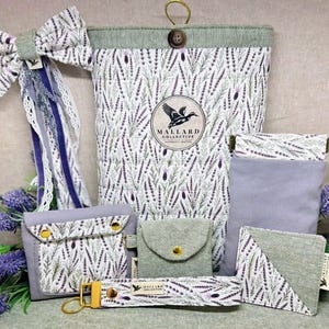 May include: A collection of fabric accessories featuring a lavender sprig pattern, including a large quilted pouch with a Mallard Collective logo, a bow hair clip, a small wallet, a coin purse, a rectangular sleeve, a corner bookmark, and a keychain.