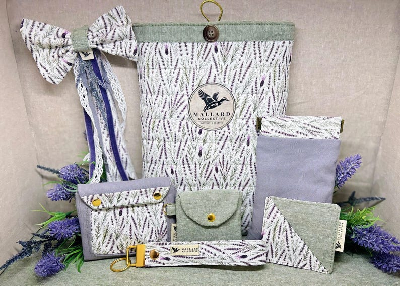 May include: A collection of fabric accessories featuring a lavender sprig pattern, including a large quilted pouch with a Mallard Collective logo, a bow hair clip, a small wallet, a coin purse, a rectangular sleeve, a corner bookmark, and a keychain.