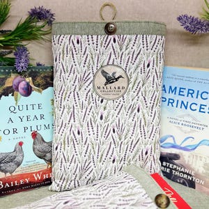 May include: A quilted fabric book sleeve featuring a lavender sprig pattern, a green top border, and a single button closure. A circular logo patch in the center displays a mallard duck silhouette and the text Mallard Collective, Crafted.