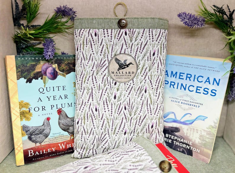 May include: A quilted fabric book sleeve featuring a lavender sprig pattern, a green top border, and a single button closure. A circular logo patch in the center displays a mallard duck silhouette and the text Mallard Collective, Crafted.